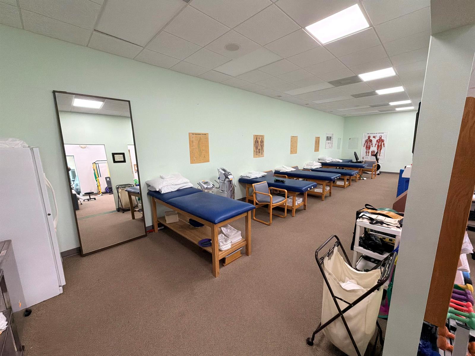 A physical therapy room features multiple treatment tables, medical posters, and a large floor mirror — 5055 S Congress Avenue # 304, Atlantis FL 33461, ATLANTIS, FL
