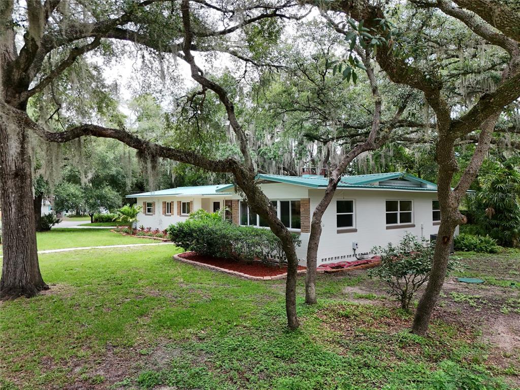 A white single-story house with a green metal roof sits among large oak trees — 22 POINSETTIA DRIVE, DELAND, FL