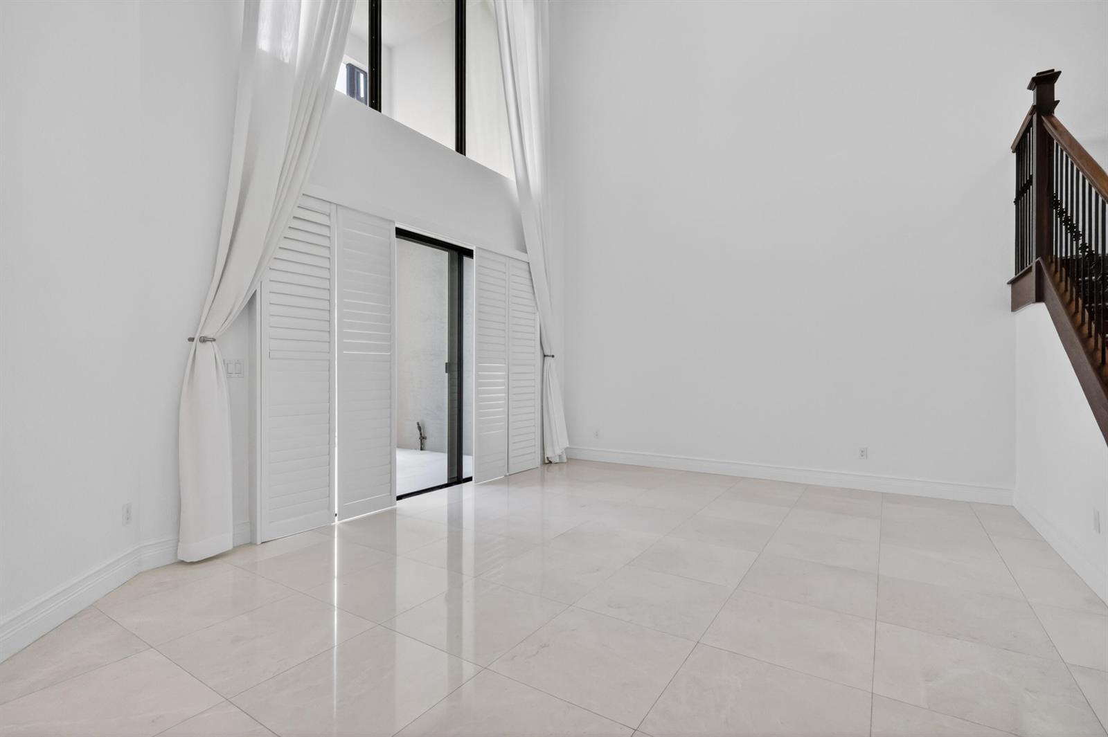 A bright, high-ceilinged room features white tile flooring, tall windows with curtains, and a staircase — 9524 Eden Roc Court, Delray Beach FL 33446, DELRAY BEACH, FL