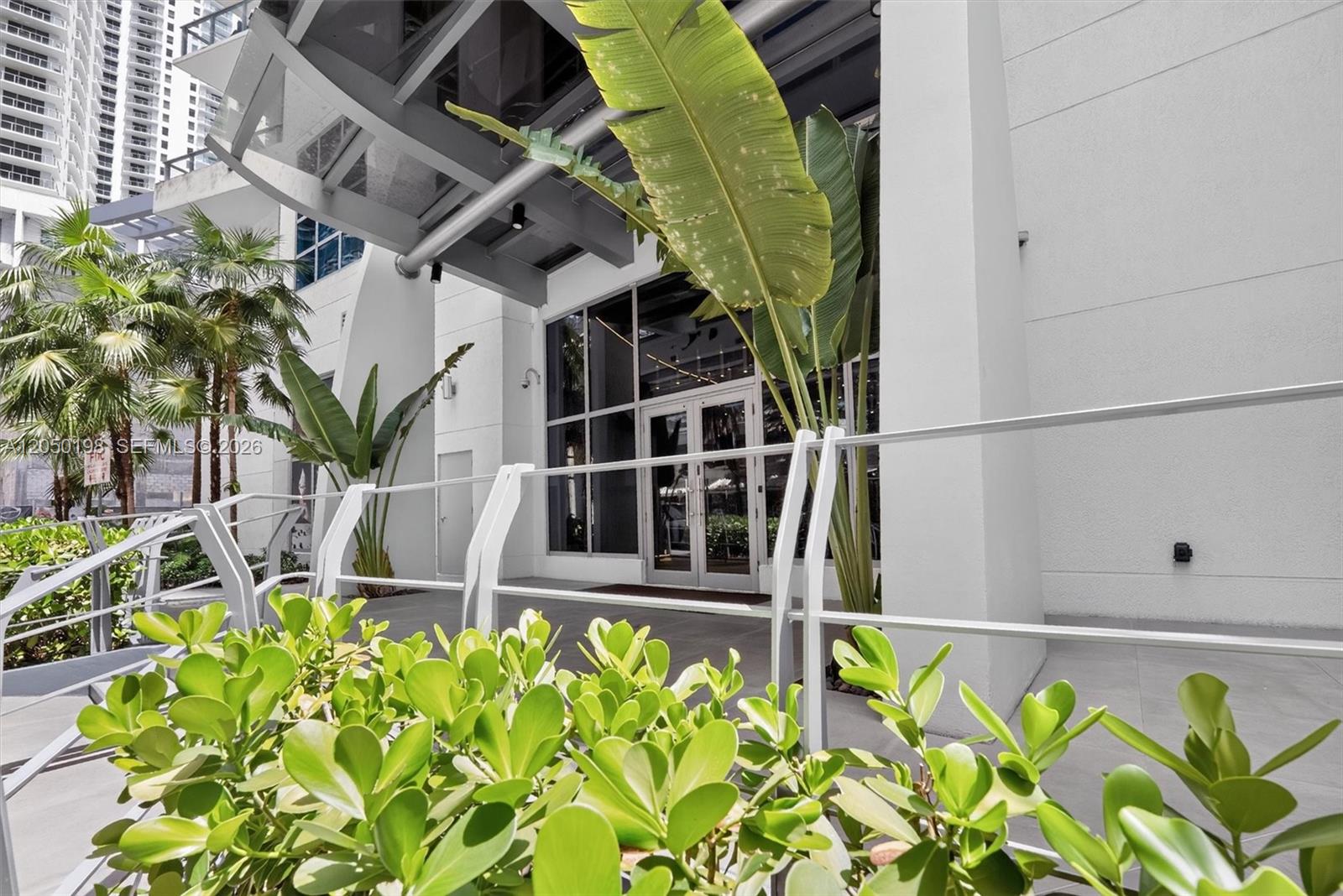 A modern building entrance features a glass door, metal railing, and large tropical plants — 1100 S Miami Ave # 3707, Miami FL 33130, MIAMI, FL