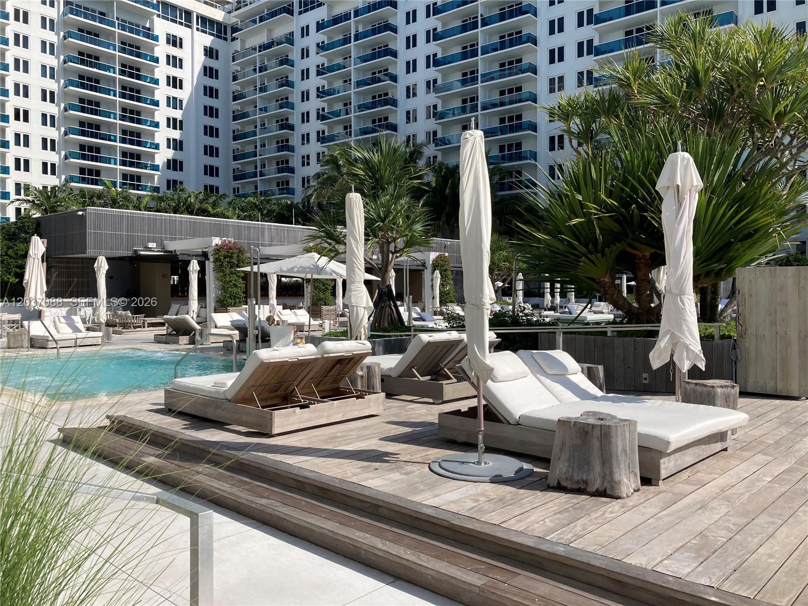 A swimming pool area features wooden lounge chairs, white umbrellas, and a large apartment building — 2301 Collins Ave # 614, Miami Beach FL 33139, MIAMI BEACH, FL