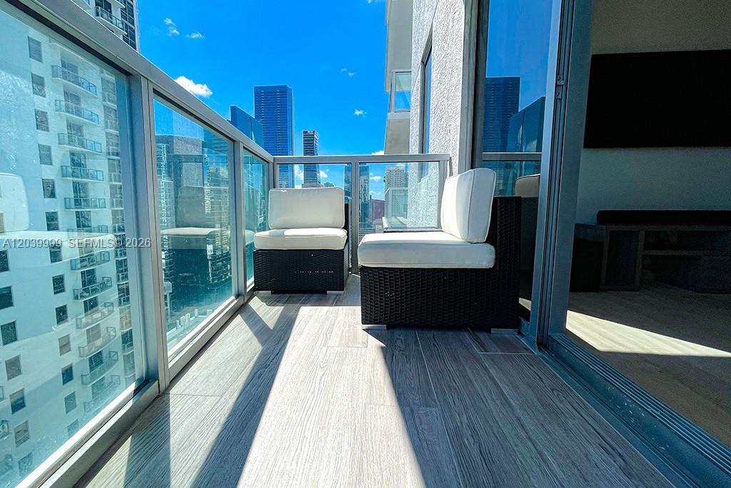 A balcony with two wicker chairs, wood-look tile flooring, and glass railings overlooking city buildings — 1050 Brickell Ave # 2208, Miami FL 33131, MIAMI, FL