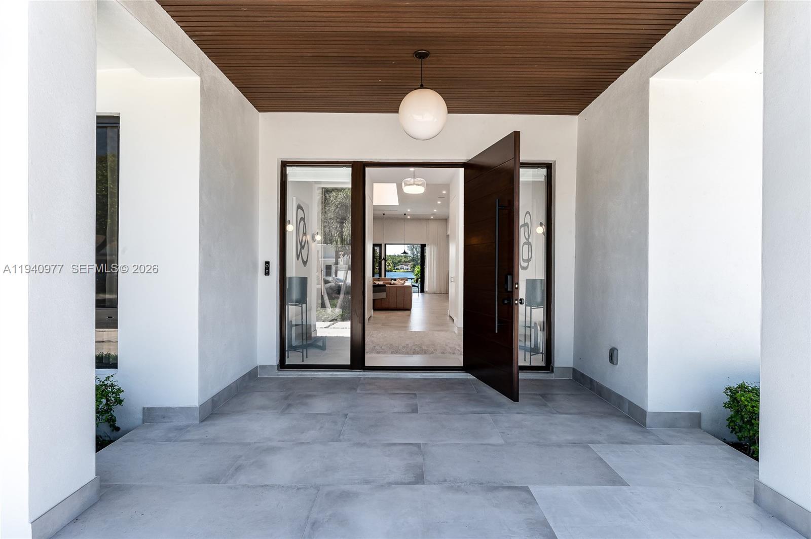 A modern entryway features a large wooden pivot door, a globe pendant light, and tiled flooring — 2560 NE 199th St, Miami FL 33180, MIAMI, FL