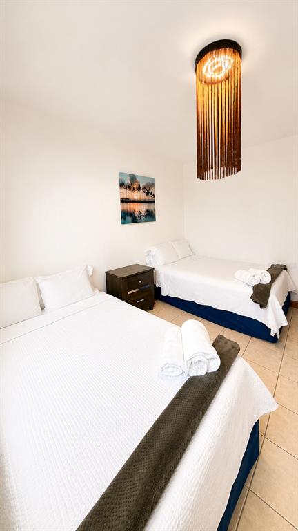 A bedroom features two beds with white linens, a nightstand, and a decorative fringe light — CII Via 2 VILLA FONTANA, CAROLINA, PR