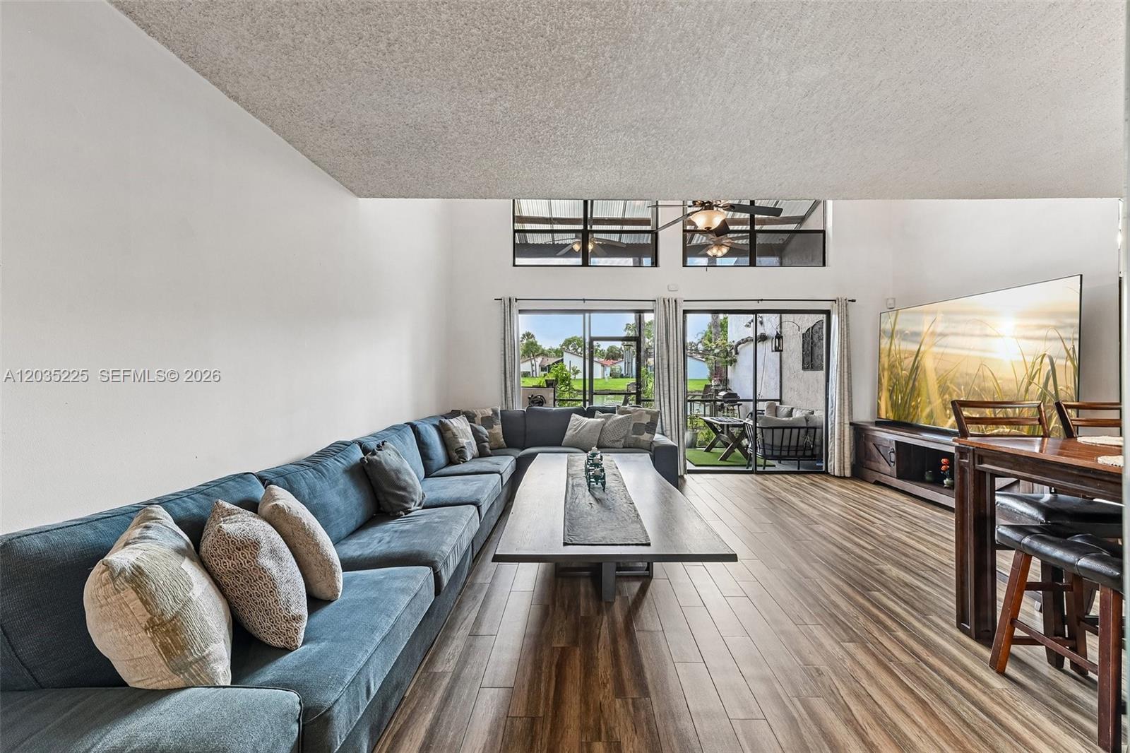 A living room features a large blue sectional sofa, wood-look flooring, and high windows — 207 NW 45th Ave, Deerfield Beach FL 33442, DEERFIELD BEACH, FL