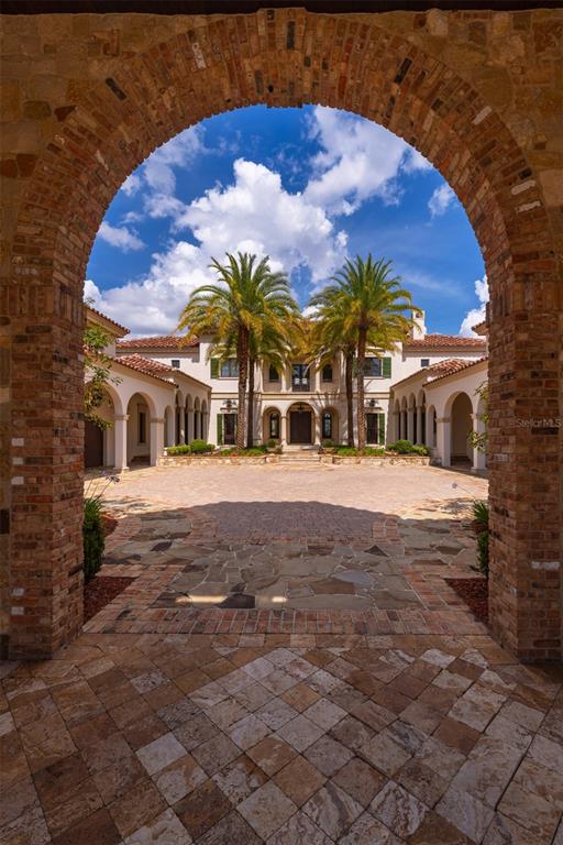 A stone archway frames a view of a Mediterranean-style mansion with palm trees and courtyard — 6119 GROSVENOR SHORE DRIVE, WINDERMERE, FL