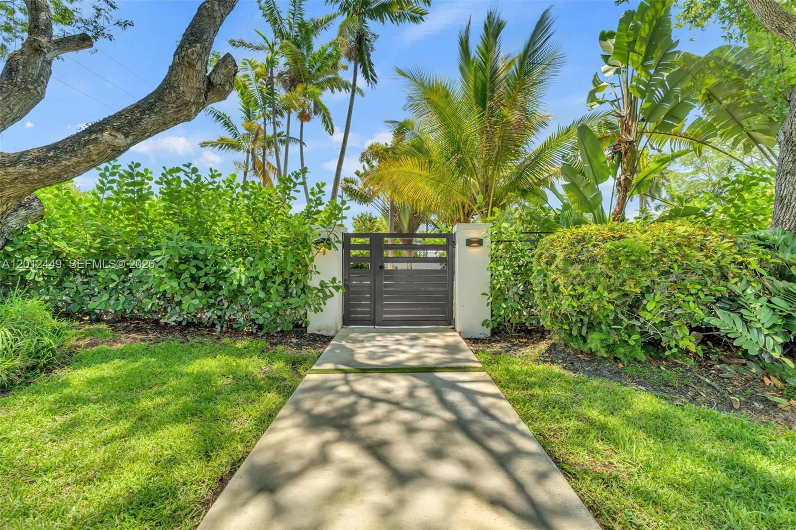 A concrete walkway leads to a dark metal gate flanked by white pillars and hedges — 6200 SW 118th St, Pinecrest FL 33156, PINECREST, FL