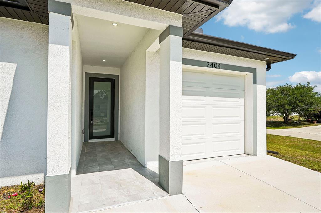 A white stucco house exterior features a covered entryway, a glass-paneled door, and a garage — 2406 HIGHTOWER AVENUE S, LEHIGH ACRES, FL