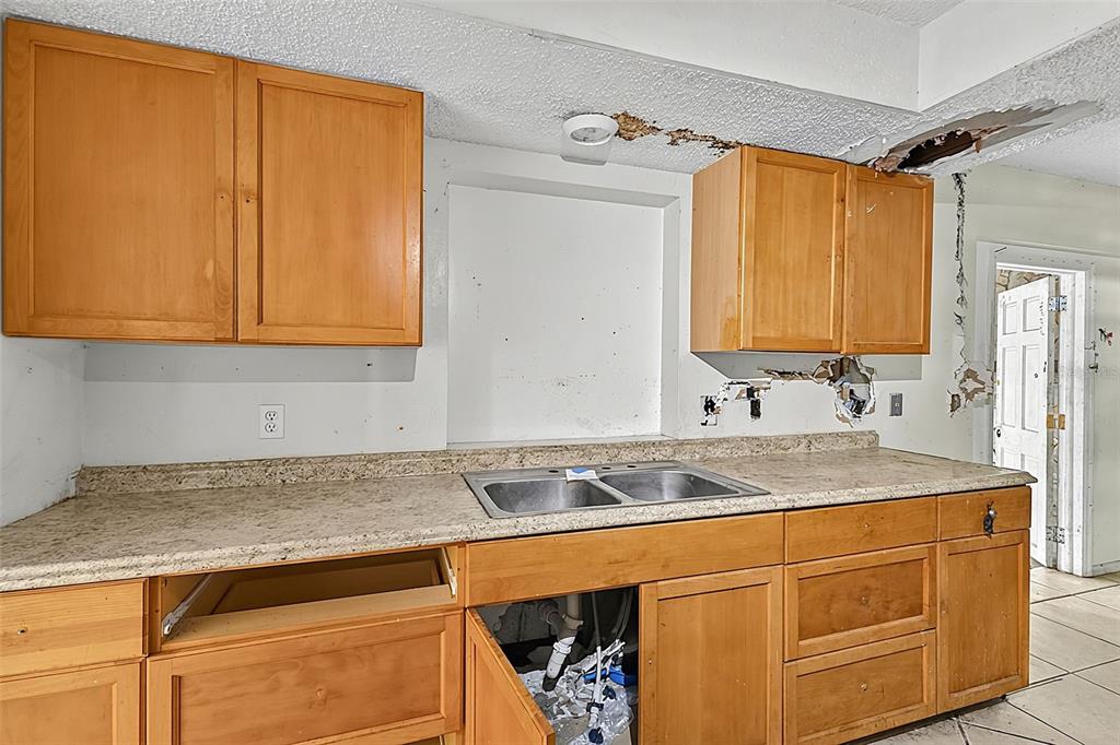 A dilapidated kitchen features wood cabinets, tile flooring, and significant damage to walls and ceilings — 4830 COCO PALM DRIVE, SEBRING, FL