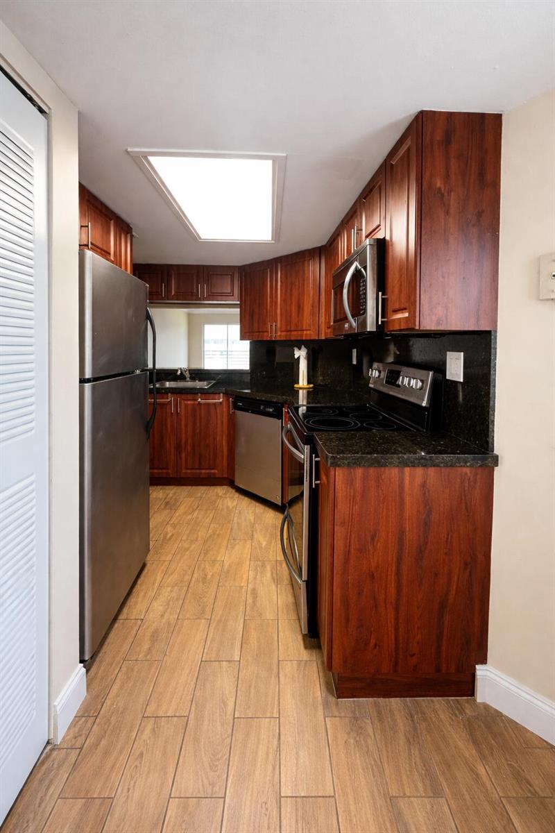 A kitchen features dark wood cabinets, stainless steel appliances, black countertops, and wood-look tile flooring — 2837 N Course Drive # 202, Pompano Beach FL 33069, POMPANO BEACH, FL