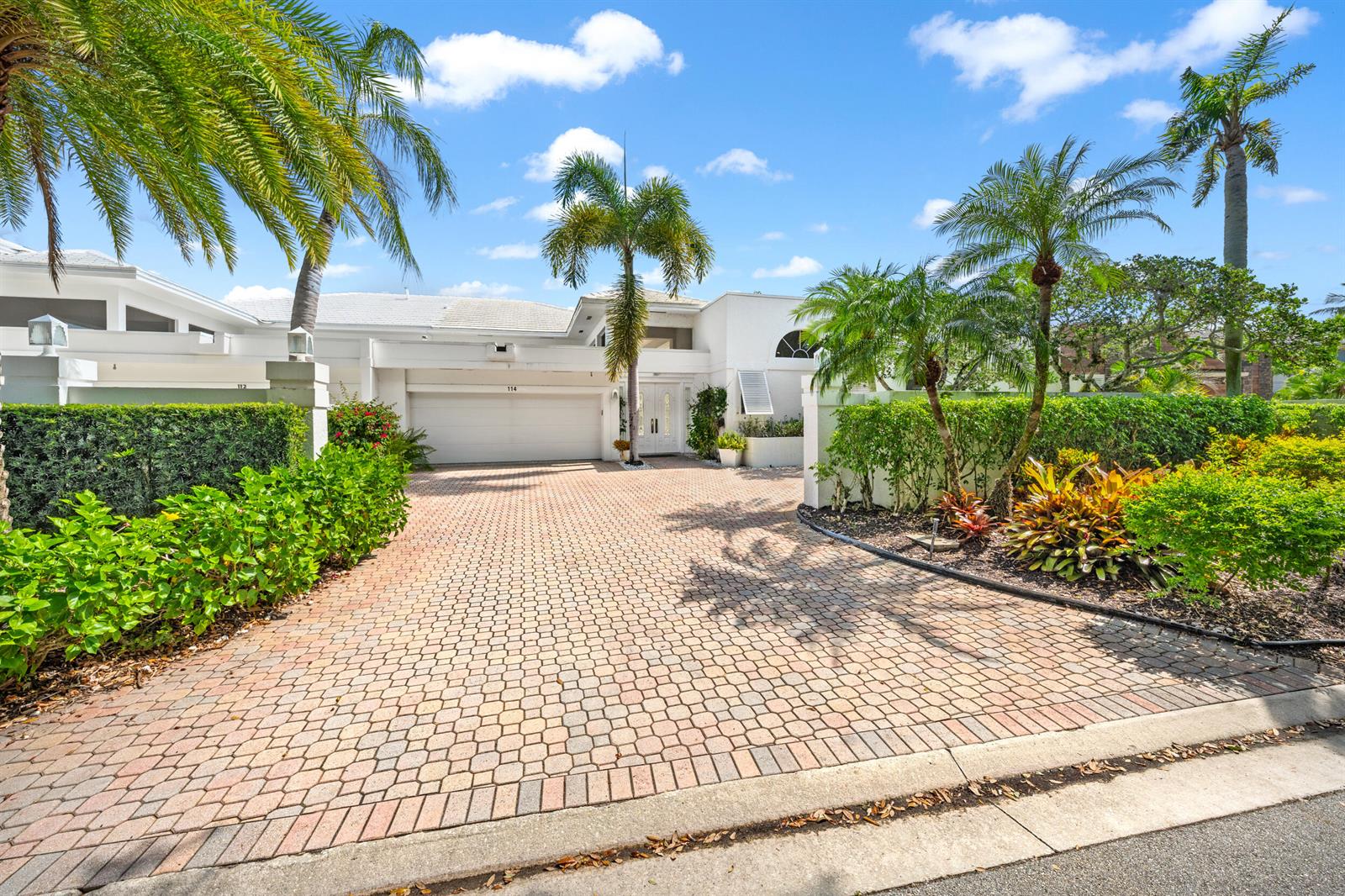 A white house with a paved driveway, palm trees, and manicured hedges under a blue sky — 114 Waters Edge Drive, Jupiter FL 33477, JUPITER, FL