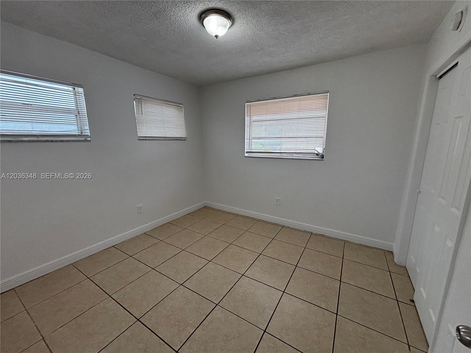 An empty room features white walls, tiled flooring, three windows with blinds, and a door — 8287 NE Miami Ct # 201, Miami FL 33138, MIAMI, FL