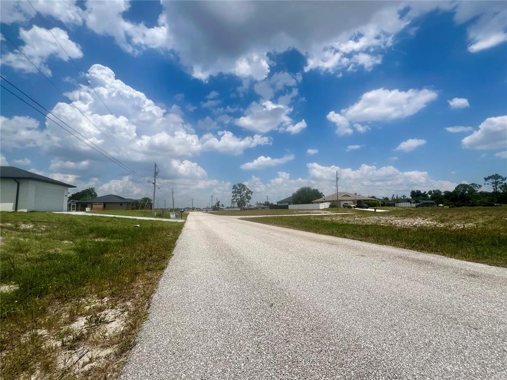 A paved road stretches through a residential neighborhood under a bright, cloudy blue sky — 1623 NW 26TH STREET, CAPE CORAL, FL