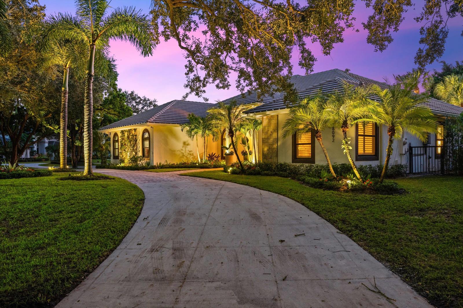 A single-story house with a curved driveway, palm trees, and landscape lighting at dusk — 18092 SE Laurel Leaf Lane, Jupiter FL 33469, JUPITER, FL