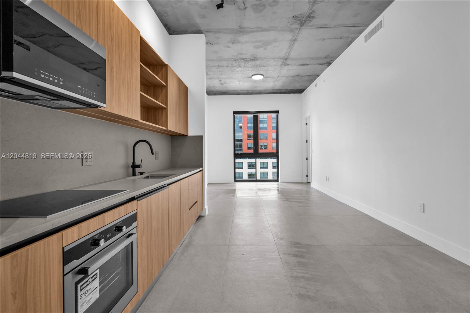 A modern kitchen with light wood cabinets and concrete ceilings leads to a windowed living area — 91 NW 27th St # 204, Miami FL 33127, MIAMI, FL