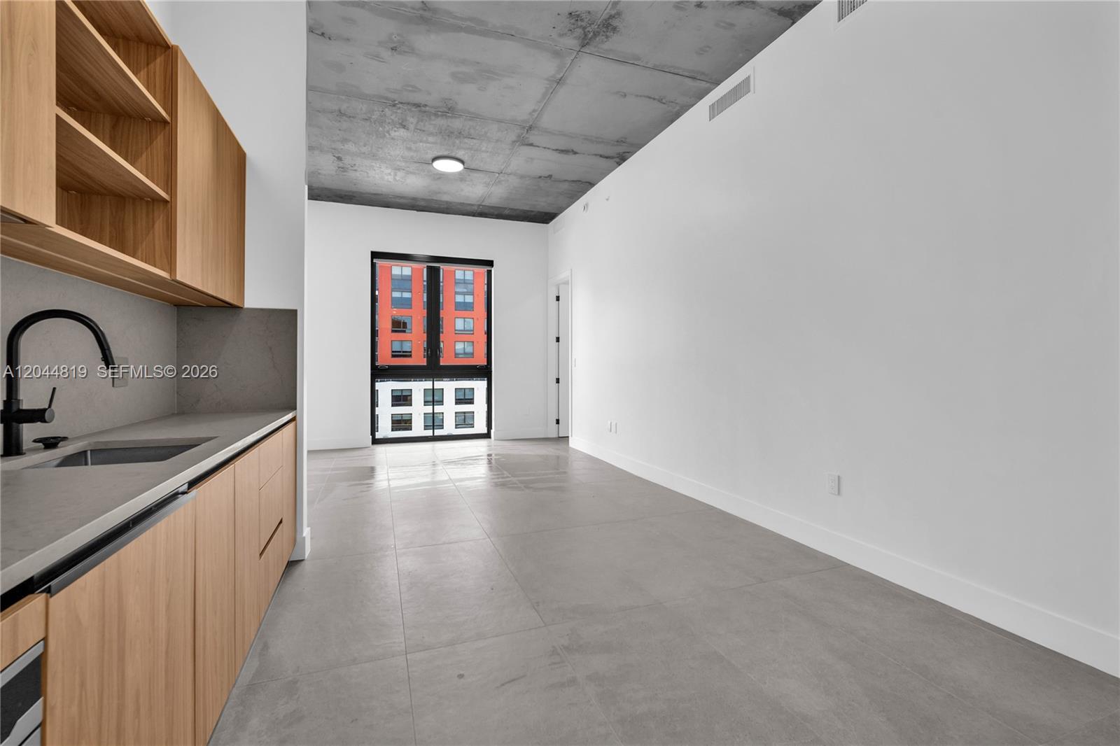 A modern kitchen with wood cabinets and concrete ceilings opens into a bright living area — 91 NW 27th St # 204, Miami FL 33127, MIAMI, FL