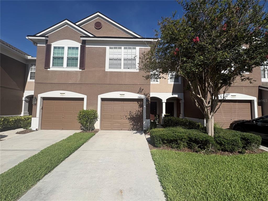 A two-story townhouse exterior features brown stucco walls, three garage doors, and a front lawn — 4605 BARNSTEAD DRIVE, RIVERVIEW, FL