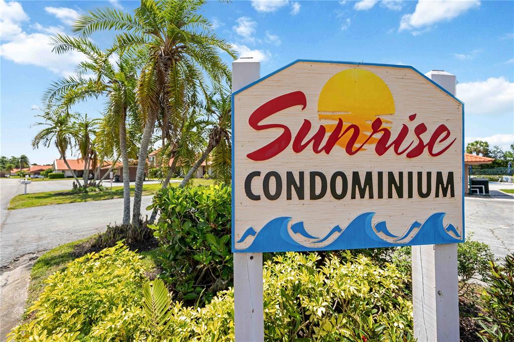 A sign for Sunrise Condominium stands outdoors near a palm tree and paved road — 6507 DRAW LANE UNIT 68, SARASOTA, FL