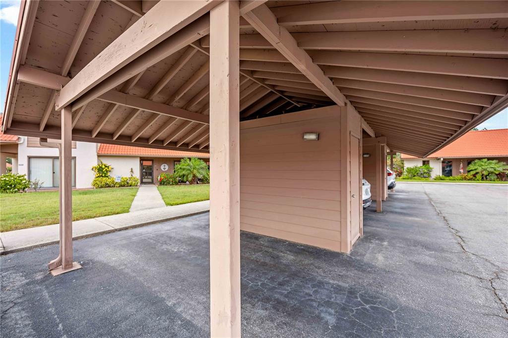A covered carport structure with wooden beams and a central storage unit in a parking lot — 6507 DRAW LANE UNIT 68, SARASOTA, FL