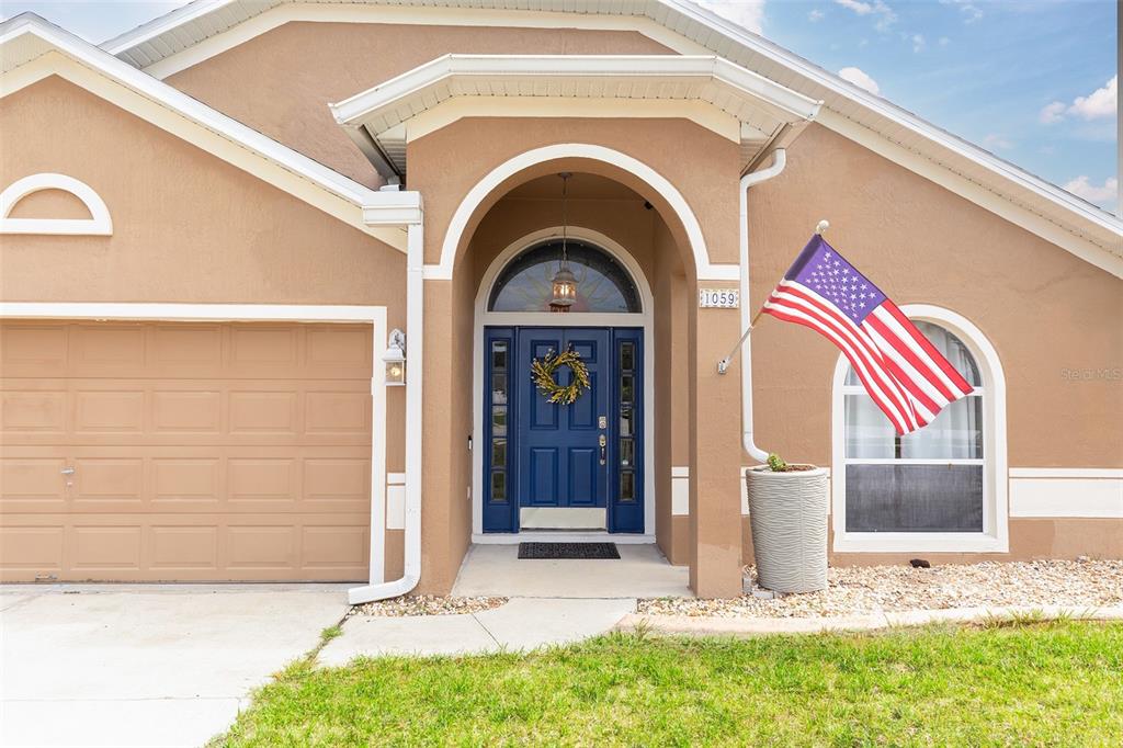 A tan house exterior features a blue front door, an arched entryway, and an American flag — 1059 BLUEGRASS DRIVE, GROVELAND, FL