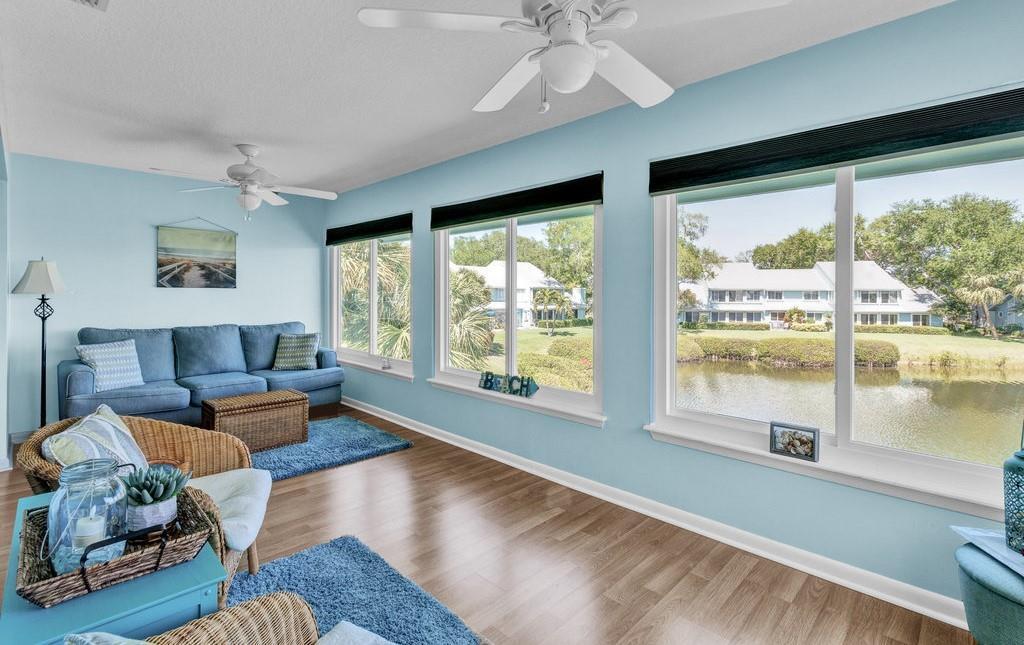 A living room with light blue walls, wood-look flooring, and large windows overlooking a pond — 6408 S River Run Drive, Sebastian FL 32958, SEBASTIAN, FL