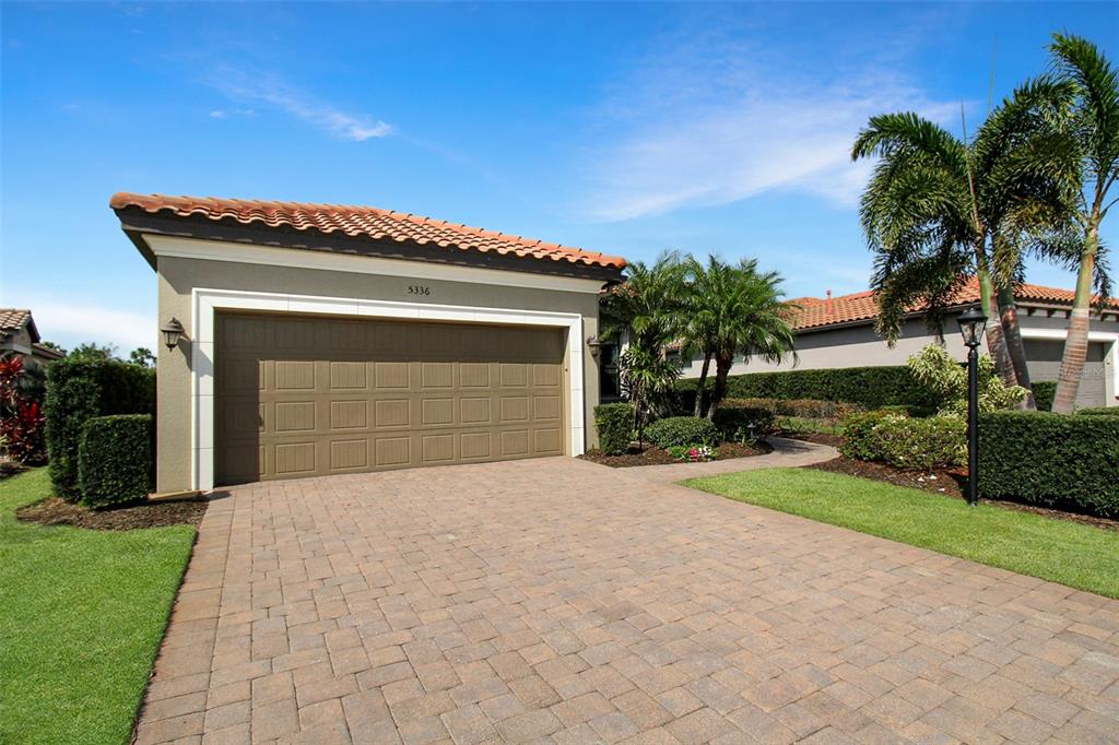 A beige house exterior features a brown garage door, a paver driveway, and palm trees — 5336 VACCARO COURT, BRADENTON, FL