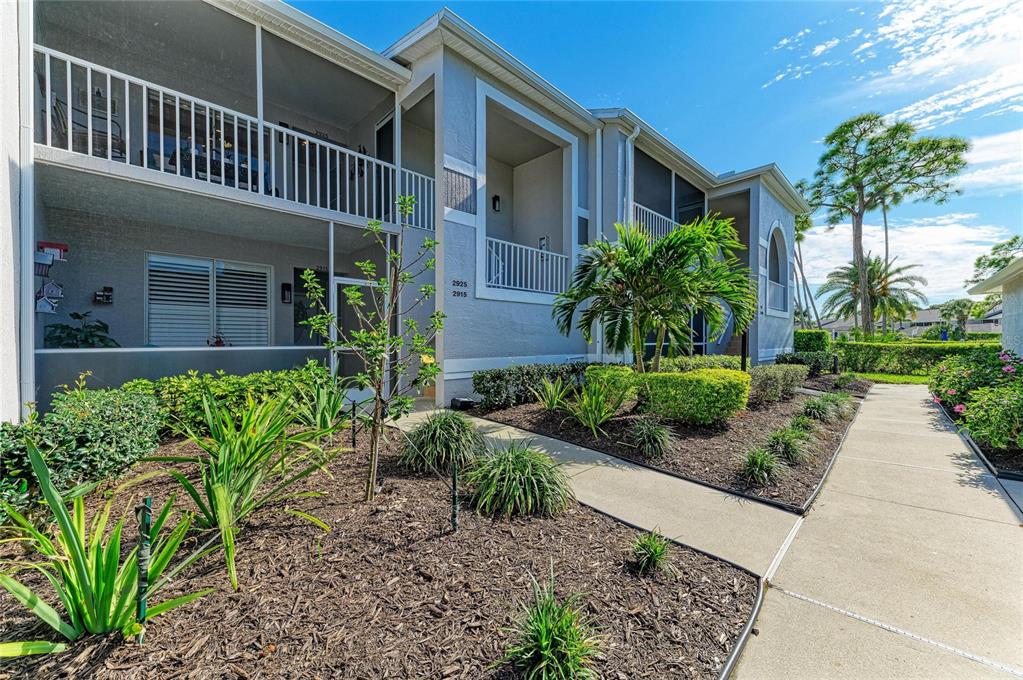 A two-story apartment building exterior features screened balconies, a concrete walkway, and landscaped garden beds — 4639 CHAPEL HILL DRIVE UNIT 2925, SARASOTA, FL