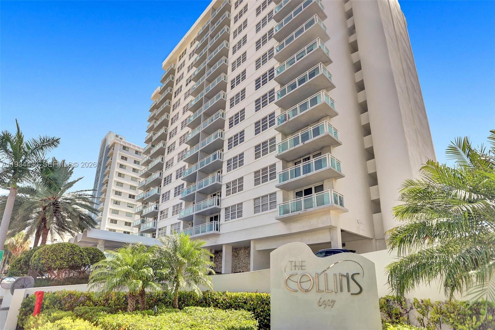 A tall white residential building with balconies stands behind a sign reading The Collins — 6917 Collins Ave # 503, Miami Beach FL 33141, MIAMI BEACH, FL