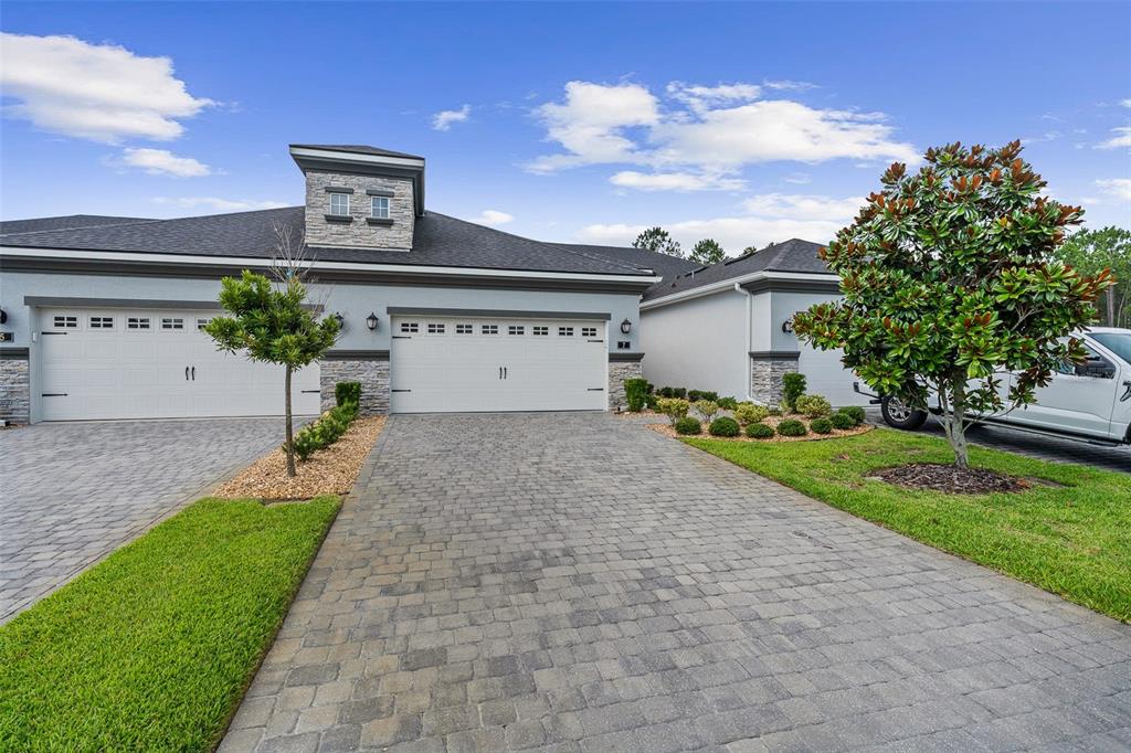A light-colored house features a stone-accented tower, two white garage doors, and a paver driveway — 7 NEWHAVEN LANE, ORMOND BEACH, FL