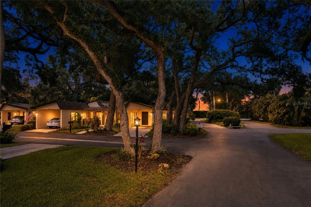 A low-slung residential building with carports stands under large trees at twilight — 6947 WOODWIND DRIVE UNIT 77, SARASOTA, FL