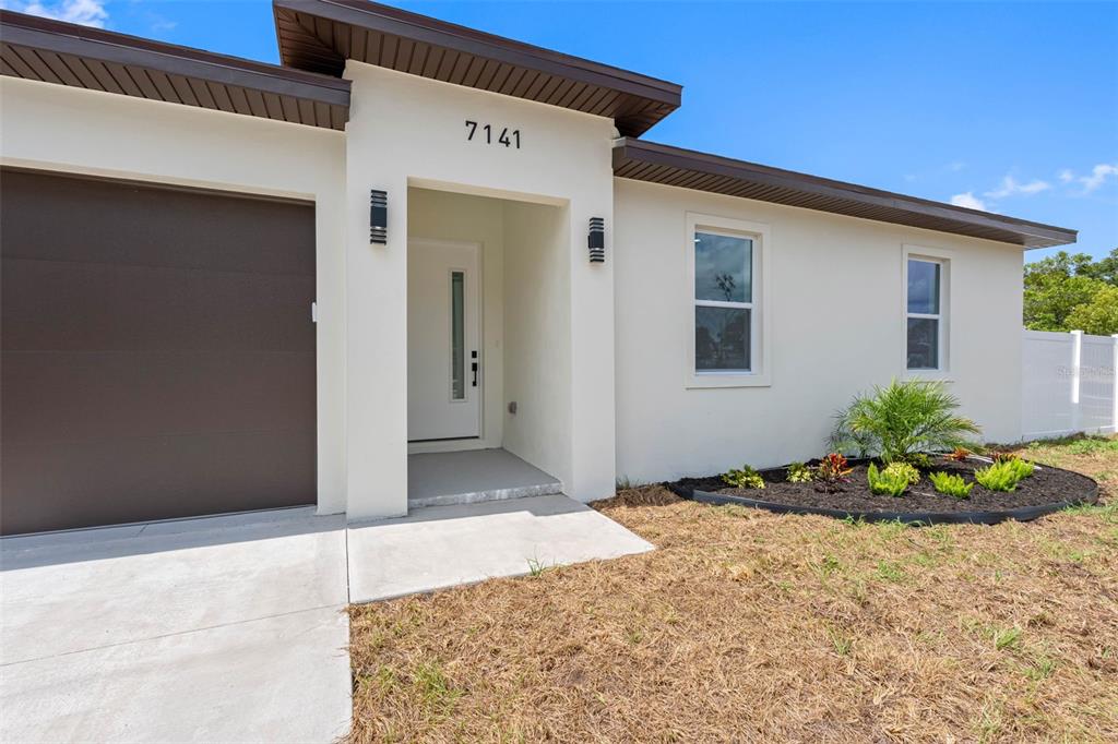 The exterior of a modern house features a white stucco facade, brown garage, and landscaping — 7141 IVANHOE DRIVE, PORT RICHEY, FL