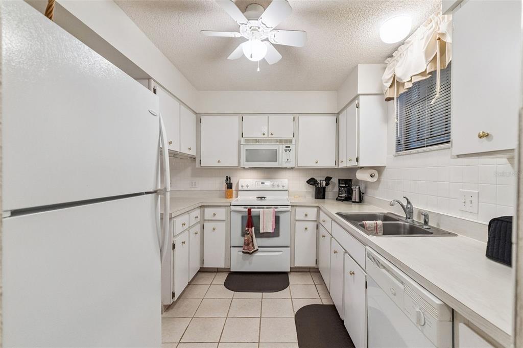 A galley-style kitchen features white cabinets, white appliances, tiled flooring, and a ceiling fan — 325 WILDER BOULEVARD UNIT 103, DAYTONA BEACH, FL