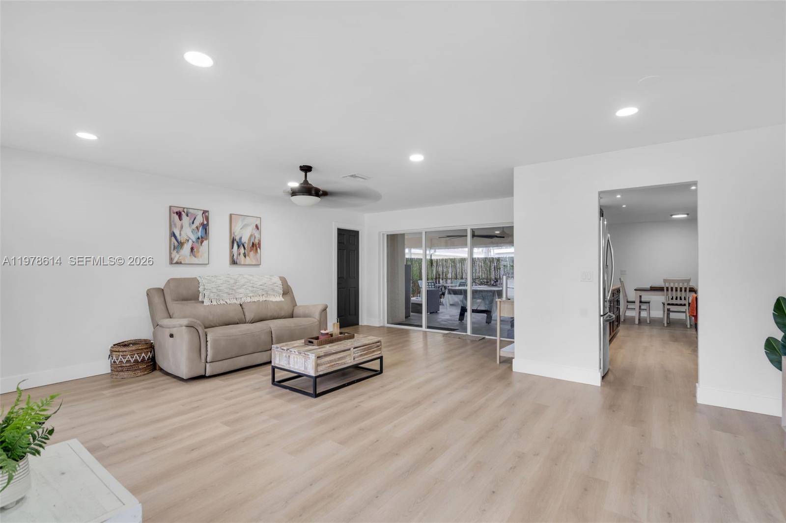 A living room features light wood flooring, a beige sofa, and sliding glass patio doors — 1180 NW 49th Ct, Deerfield Beach FL 33064, DEERFIELD BEACH, FL