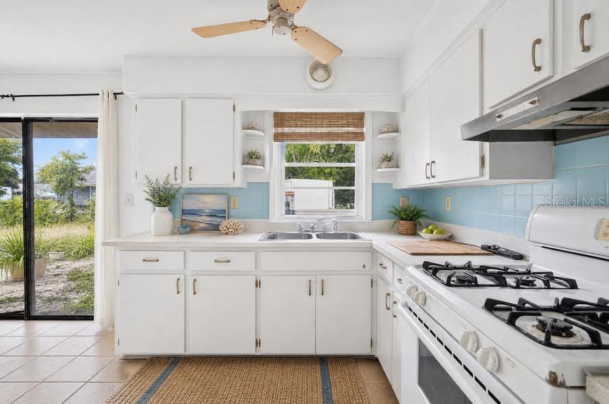 A kitchen features white cabinetry, a light blue tiled backsplash, and a window overlooking greenery — 1700 COWAN CIRCLE, TITUSVILLE, FL
