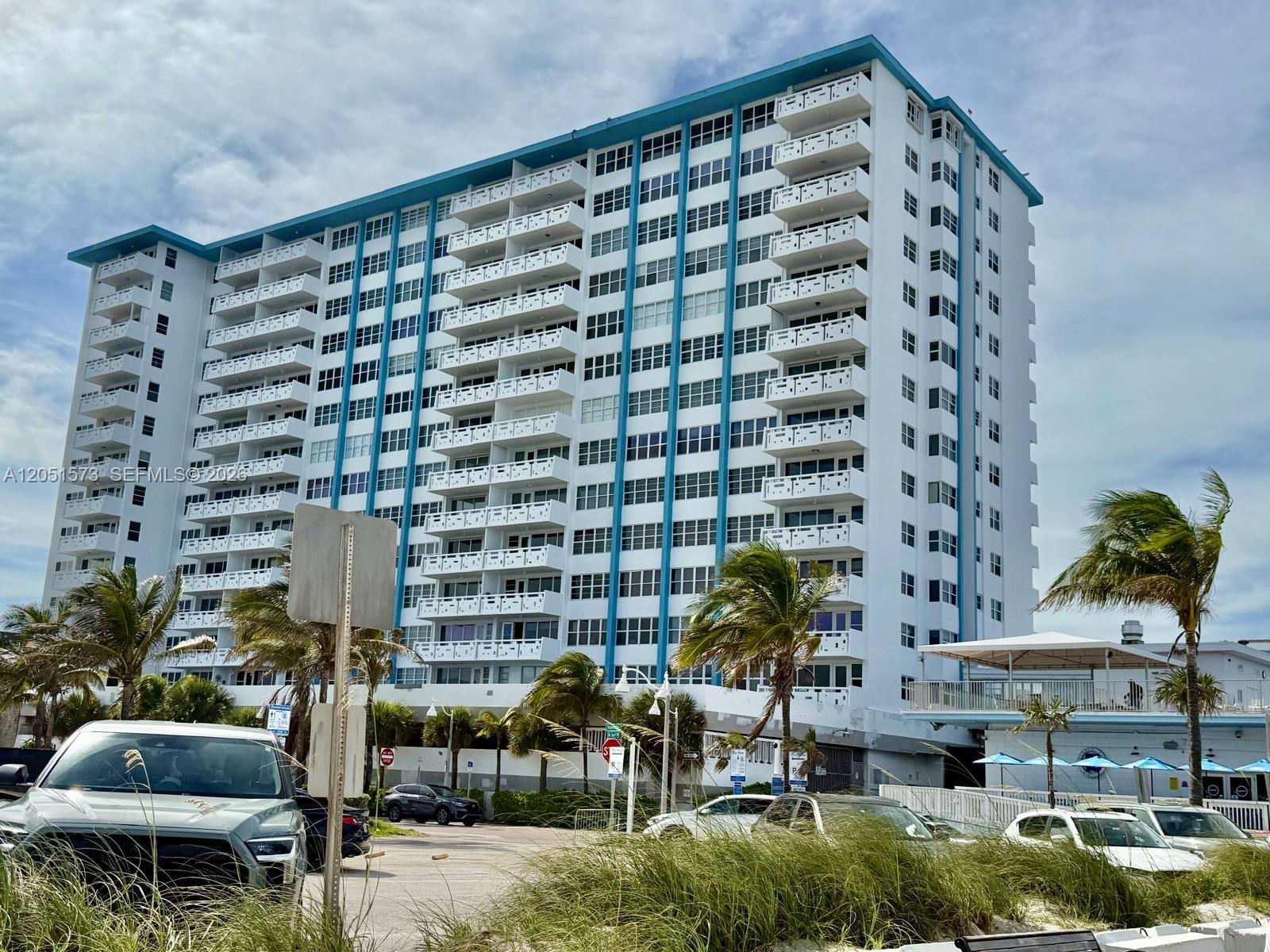 A tall, white residential building with blue accents stands behind a parking lot and palm trees — 3111 N Ocean Dr # 401, Hollywood FL 33019, HOLLYWOOD, FL