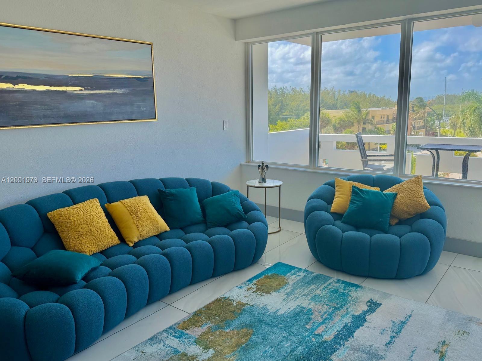 A living room features a blue tufted sofa and armchair with yellow pillows near windows — 3111 N Ocean Dr # 401, Hollywood FL 33019, HOLLYWOOD, FL