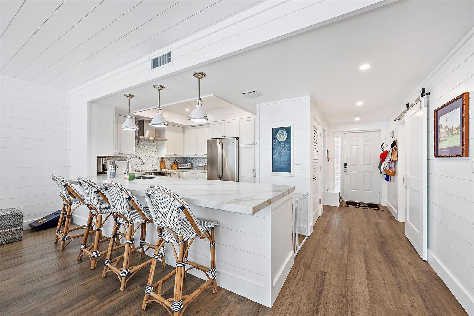 A white kitchen features a marble-topped island with four woven stools and wood flooring — 70 Celestial Way # 208, Juno Beach FL 33408, JUNO BEACH, FL