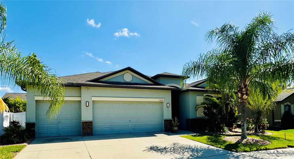 A single-story house with a three-car garage, light-colored stucco walls, and palm trees — 11604 SUNBURST MARBLE ROAD, RIVERVIEW, FL