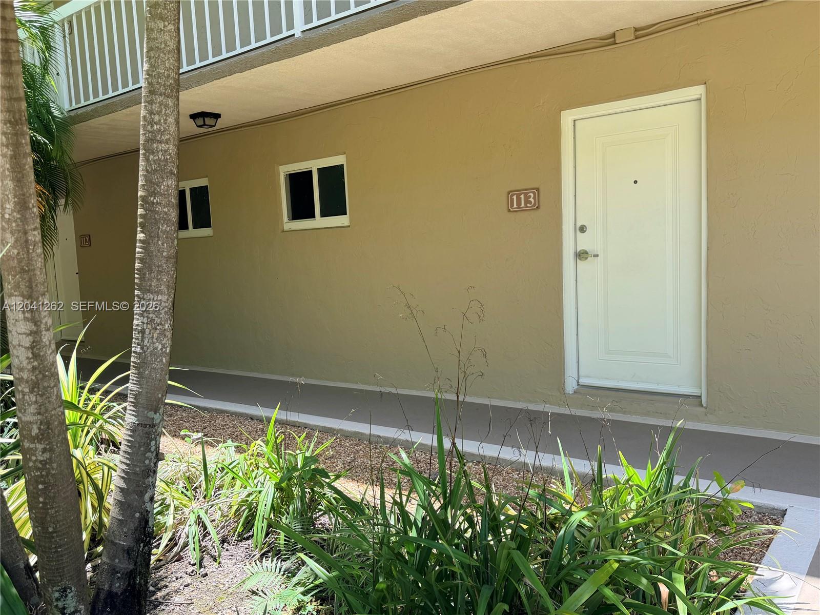 An exterior view shows a tan building wall with a white door numbered 113 — 7730 Camino Real # F113, Miami FL 33143, MIAMI, FL