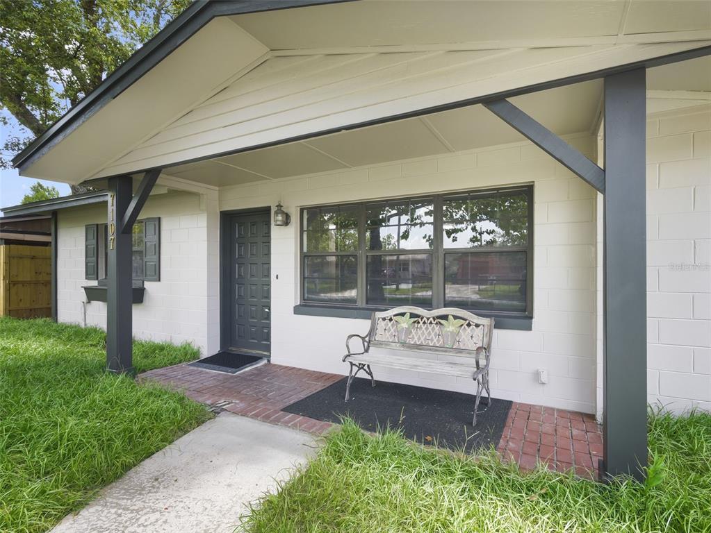 A white concrete block house exterior features a covered brick porch with a wooden bench — 7107 LEIGHTON WAY, ORLANDO, FL