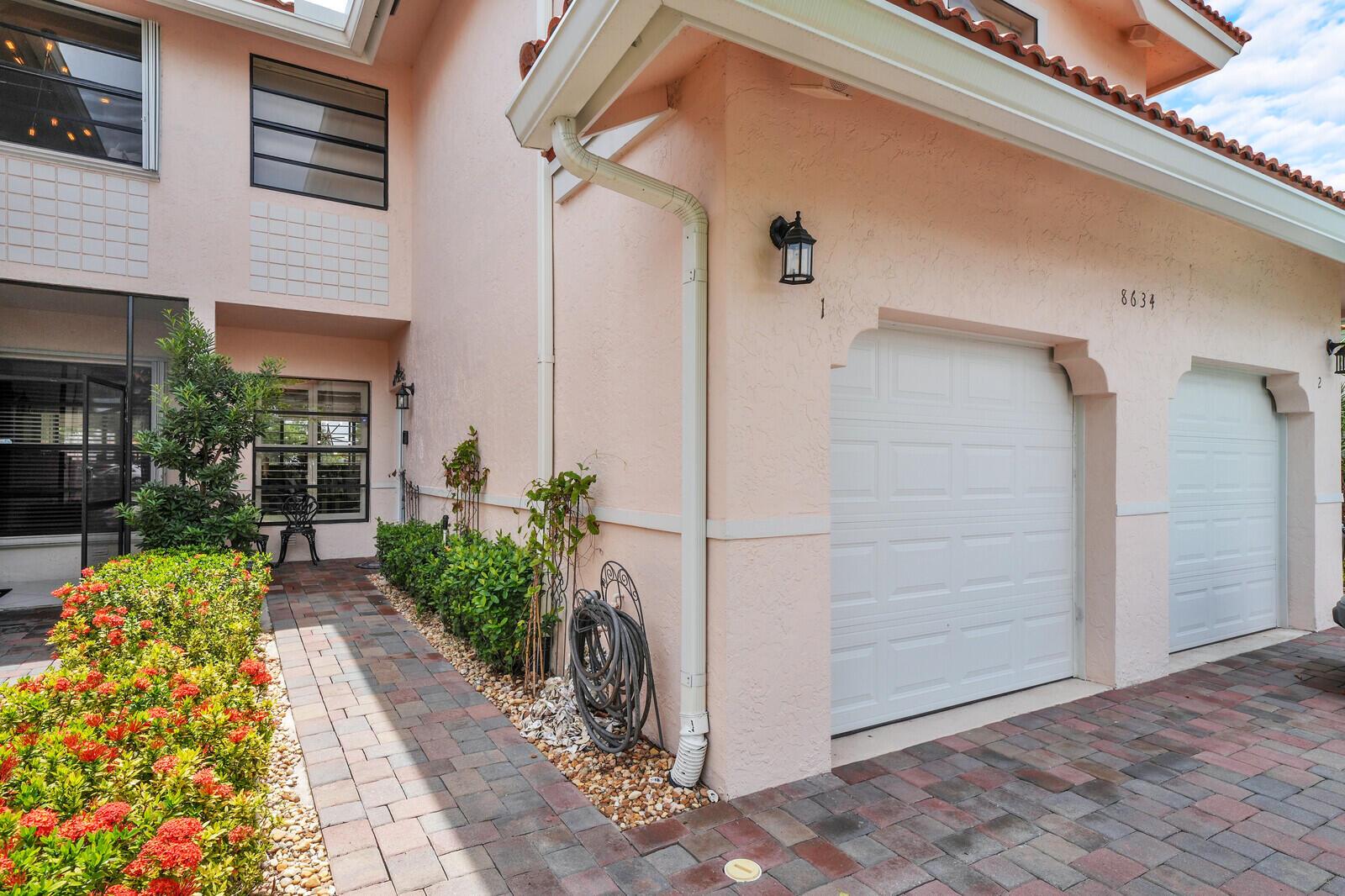 A pink stucco house exterior features a brick walkway, two white garage doors, and landscaping — 8634 Vía Reale # 1, FL 33496, FL