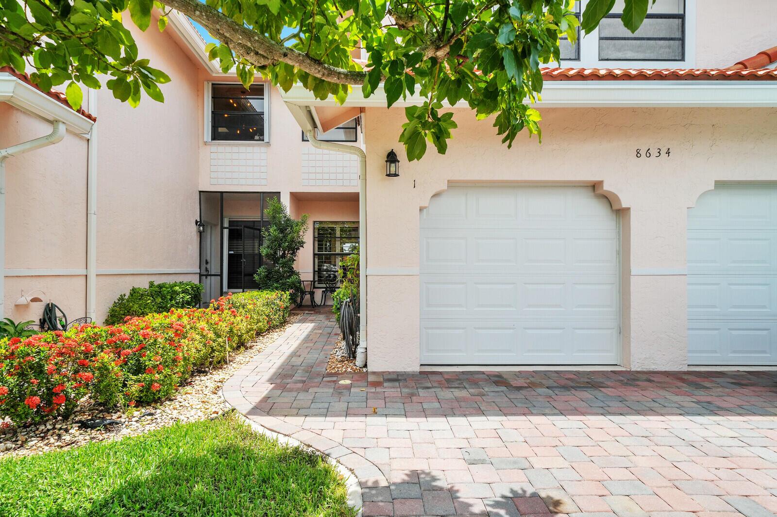 A pink stucco townhouse exterior features a paver driveway, two garage doors, and landscaped bushes — 8634 Vía Reale # 1, FL 33496, FL