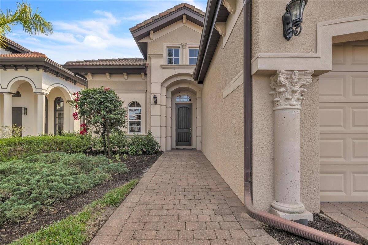 A paved walkway leads to the front entrance of a beige stucco house with columns — 7793 Ashton Road, Naples FL 34113, NAPLES, FL
