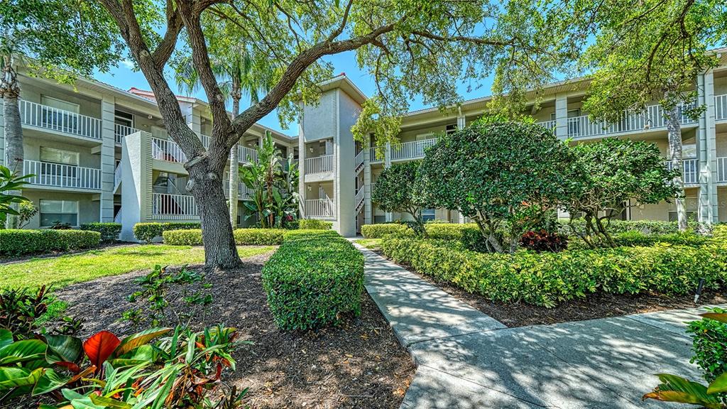 A multi-story apartment building exterior features a concrete walkway, manicured hedges, and large trees — 9330 CLUBSIDE CIRCLE UNIT 3202, SARASOTA, FL