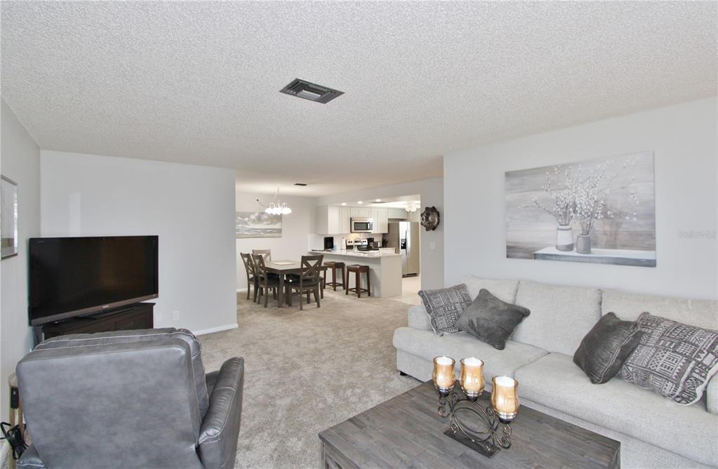 A living room with gray carpet, a sofa, a recliner, and an open-concept kitchen beyond — 9635 GREENSKEEPER DRIVE, NEW PORT RICHEY, FL