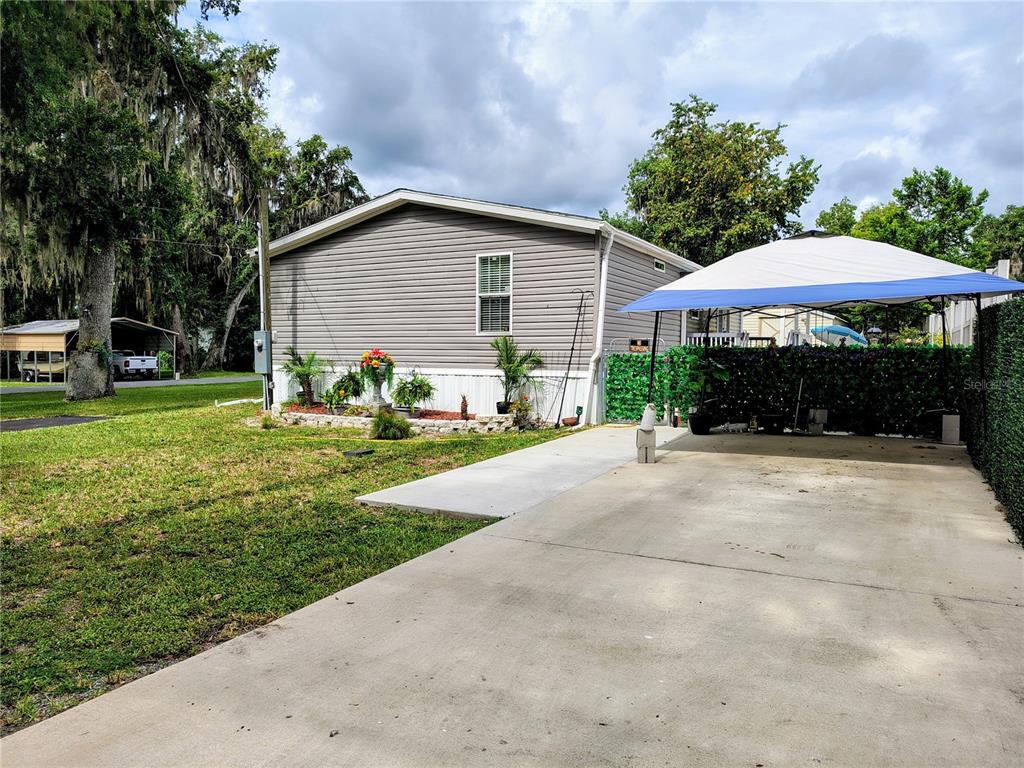 A gray mobile home features a concrete driveway, a blue canopy, and a small garden bed — 1912 CR 439C, LAKE PANASOFFKEE, FL