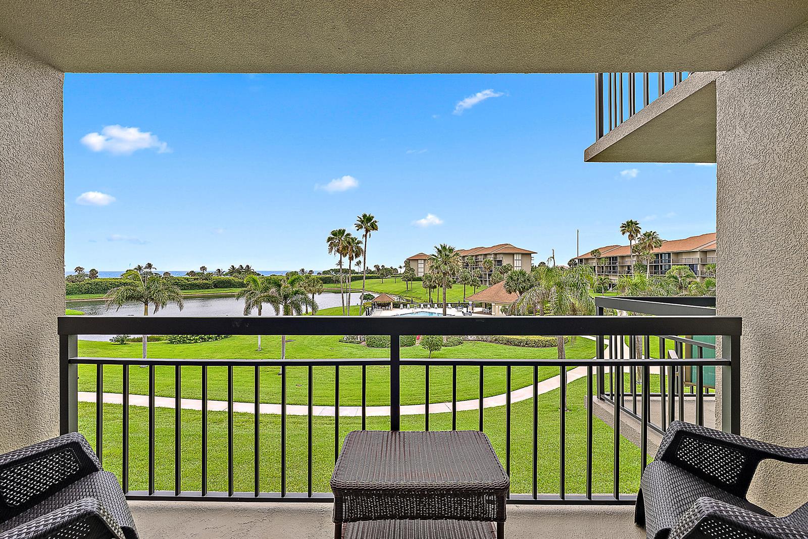 A balcony with dark wicker furniture overlooks a grassy lawn, a pond, and palm trees — 301 S Seas Drive # 305, Jupiter FL 33477, JUPITER, FL