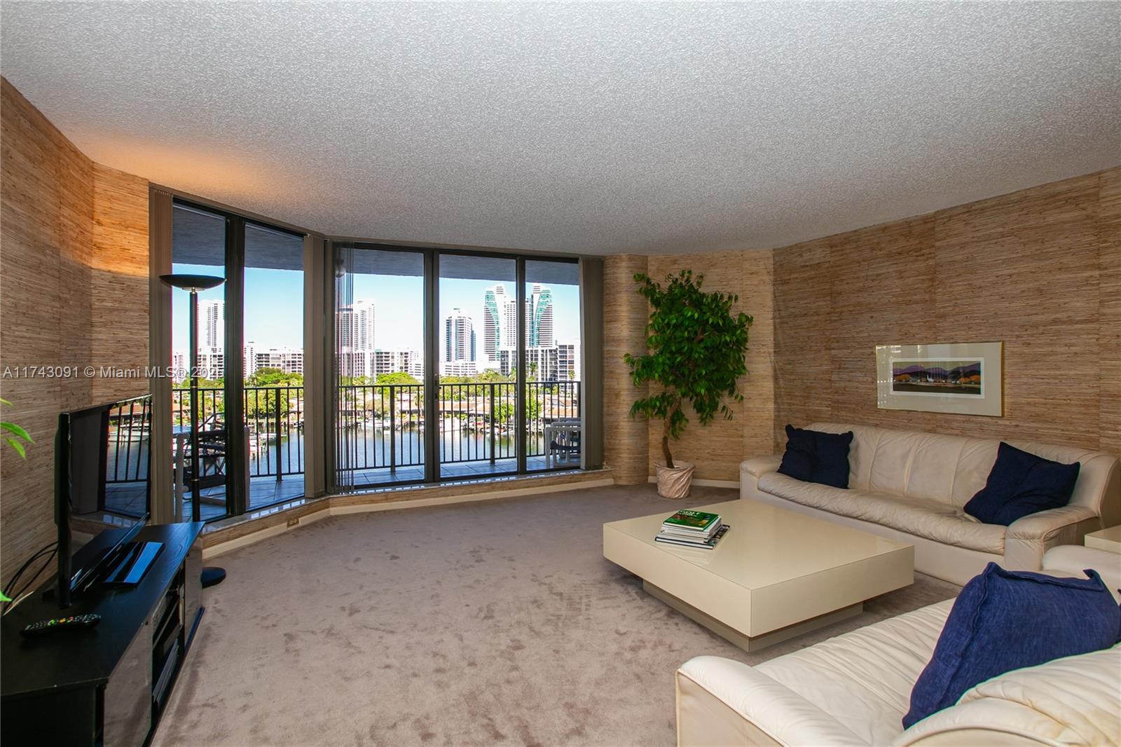 A living room features textured walls, beige carpeting, a white sofa, and floor-to-ceiling windows — 300 Three Islands Blvd # 406, Hallandale Beach FL 33009, HALLANDALE BEACH, FL