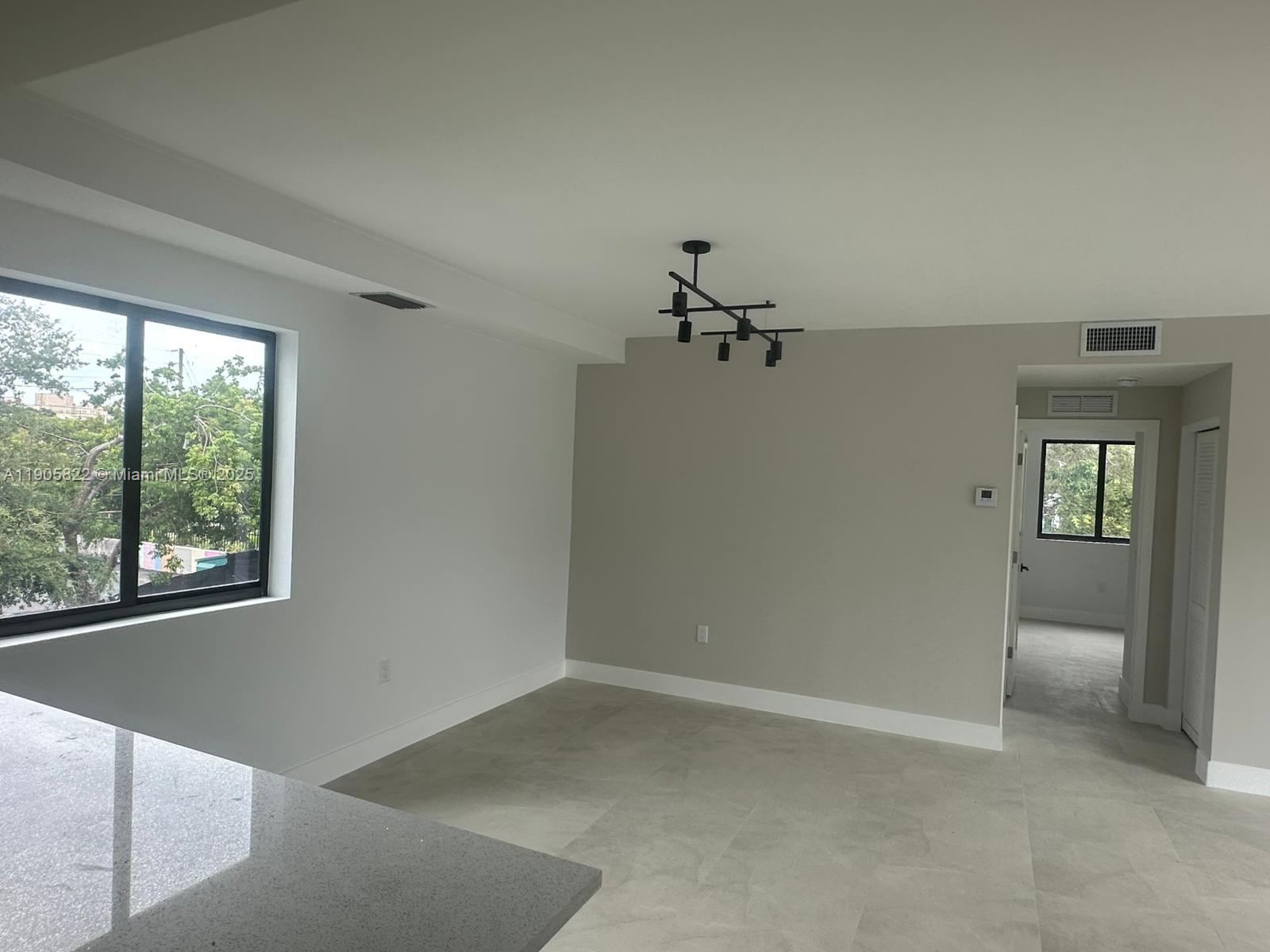 A bright, unfurnished room features light-colored tile flooring, a modern chandelier, and a large window — 5423 NE Miami Ct # 7, Miami FL 33137, MIAMI, FL