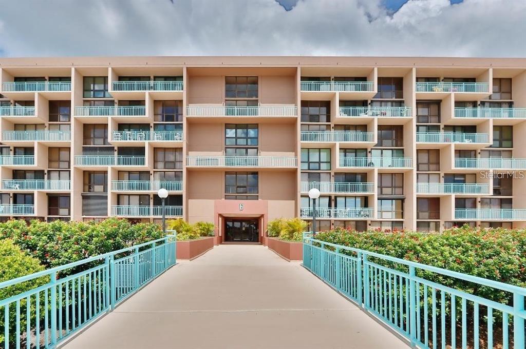 A pedestrian bridge leads to the entrance of a multi-story apartment building with balconies — 1 KEY CAPRI UNIT 302E, TREASURE ISLAND, FL